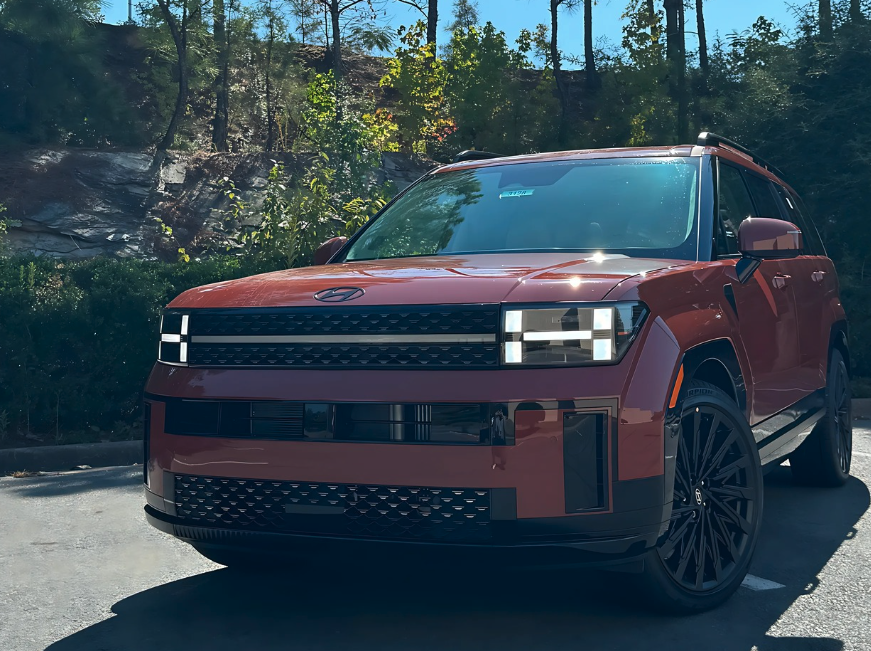 Front view of a red 2025 Hyundai Santa Fe SUV parked in a wooded area with autumn foliage near Crain Hyundai of Fort Smith.
